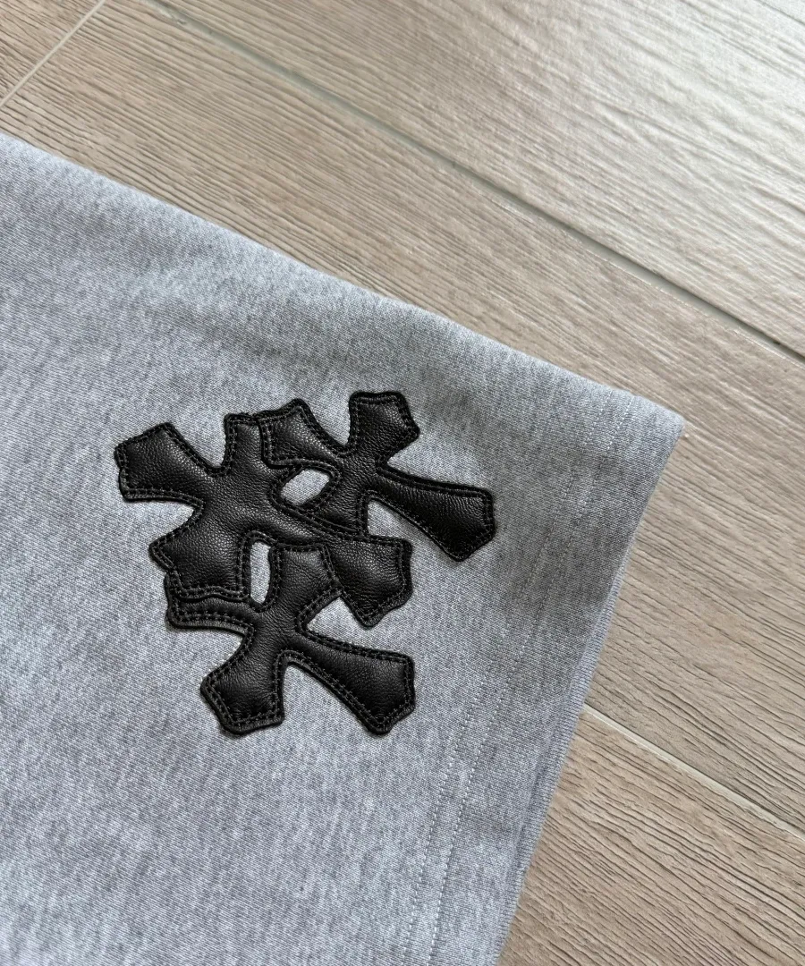 Крупный план черной кожаной нашивки-креста на серых шортах Chrome Hearts