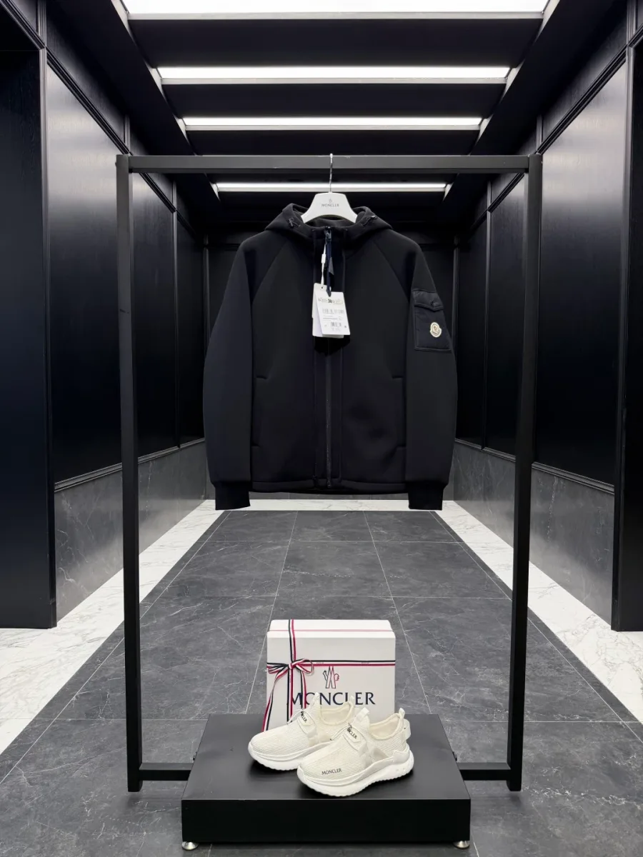 Чёрная куртка Moncler с капюшоном на вешалке в интерьере, фронтальный вид