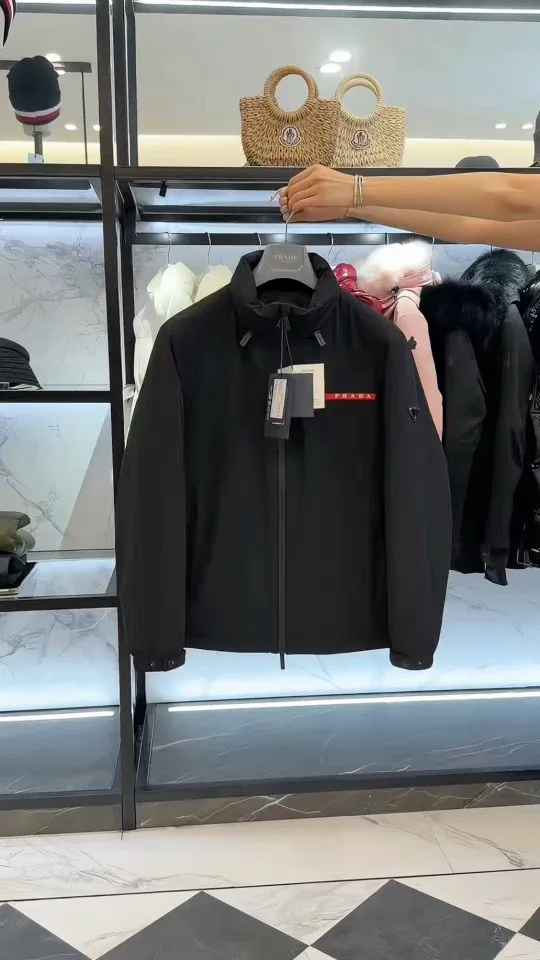 Чёрная куртка Prada на вешалке в руках, общий фронтальный вид, прямая посадка и боковые карманы