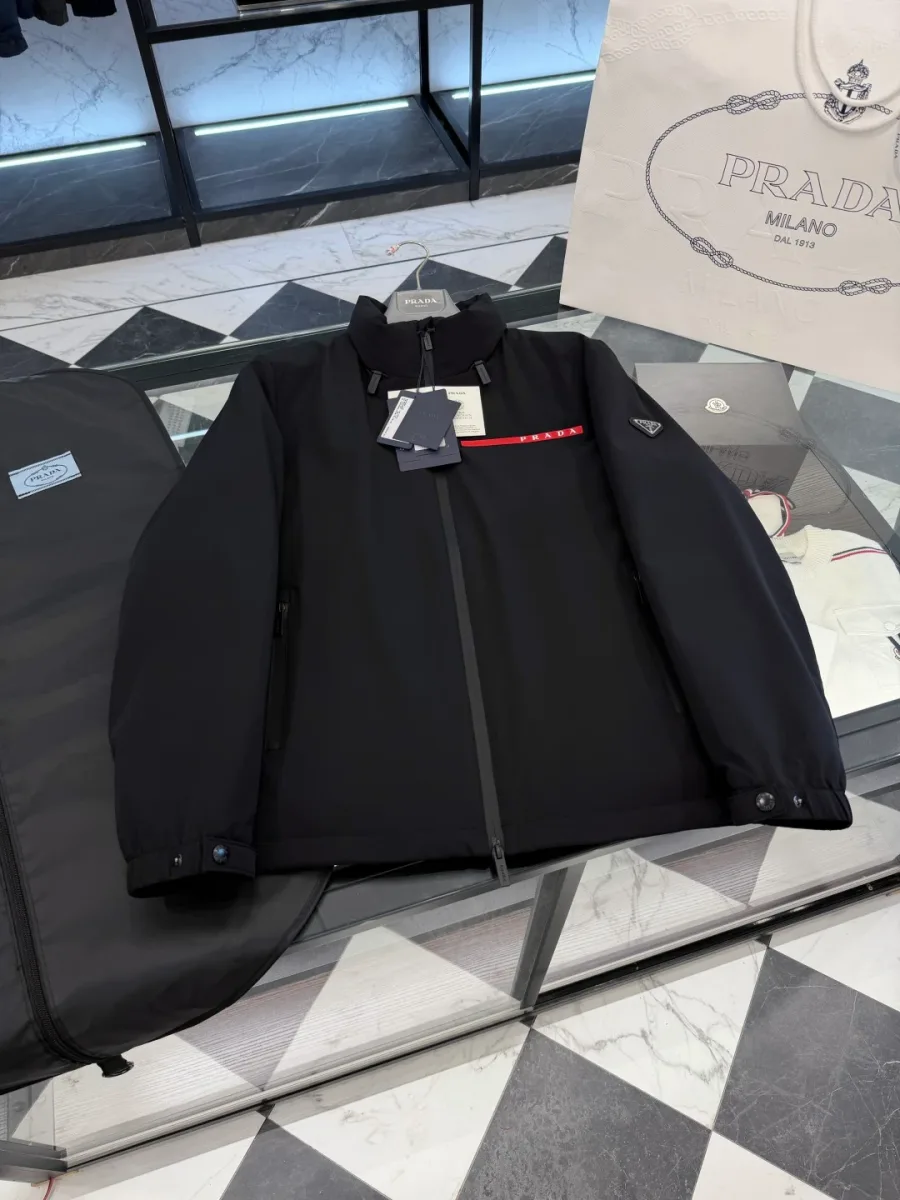 Чёрная куртка Prada в разложенном виде на витрине, ракурс сверху спереди, молния и красная планка