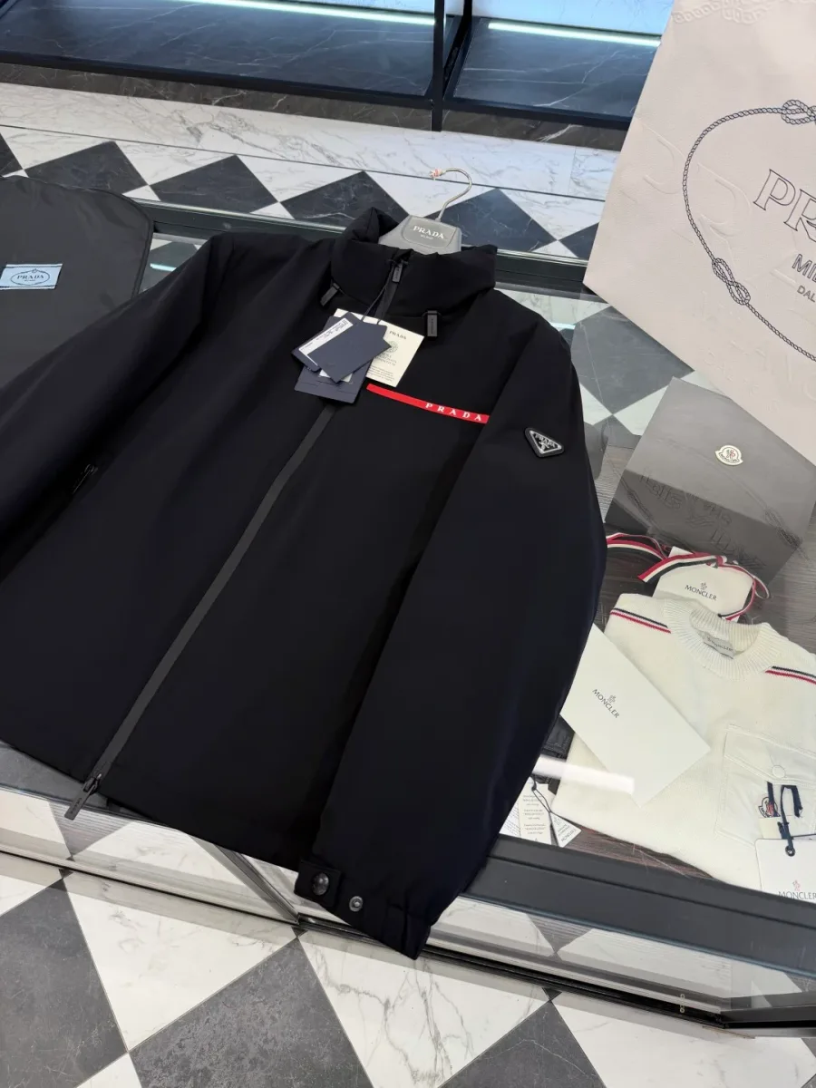 Диагональный ракурс чёрной куртки Prada: треугольный логотип на левом рукаве и манжеты на кнопках
