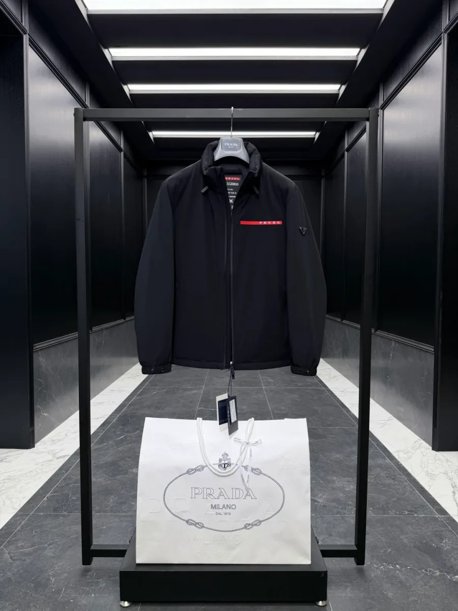 Чёрная куртка Prada с красным логотипом на груди, общий вид спереди с фирменным пакетом внизу