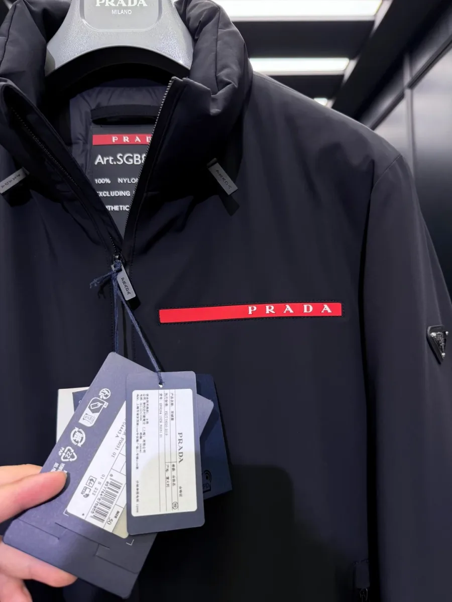 Детали воротника и груди чёрной куртки Prada: красная планка логотипа, внутренний ярлык и навесные бирки