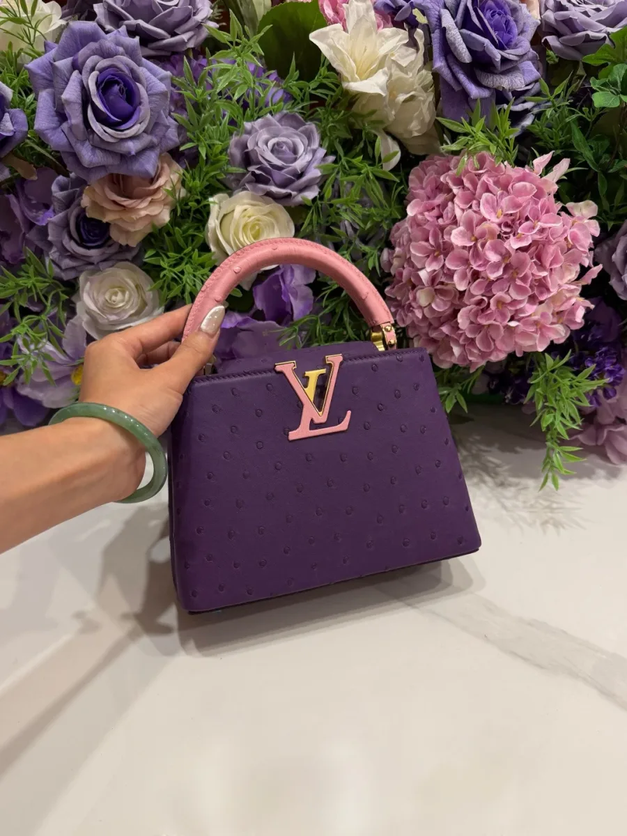 Фиолетовая сумка Capucines BB в руке на фоне цветочной композиции