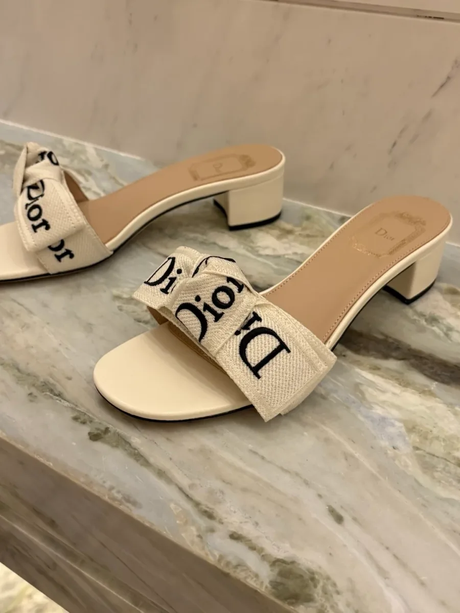Пара белых шлёпанцев на каблуке с бантом Dior на светлой поверхности, ракурс три четверти