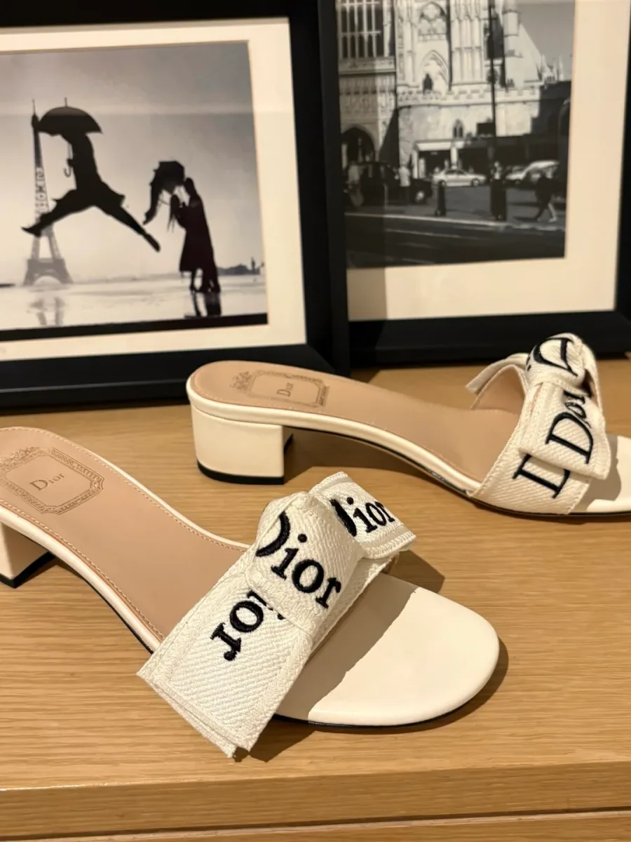 Белые шлёпанцы на каблуке с бантом Dior на фоне интерьерных картин, вид сбоку