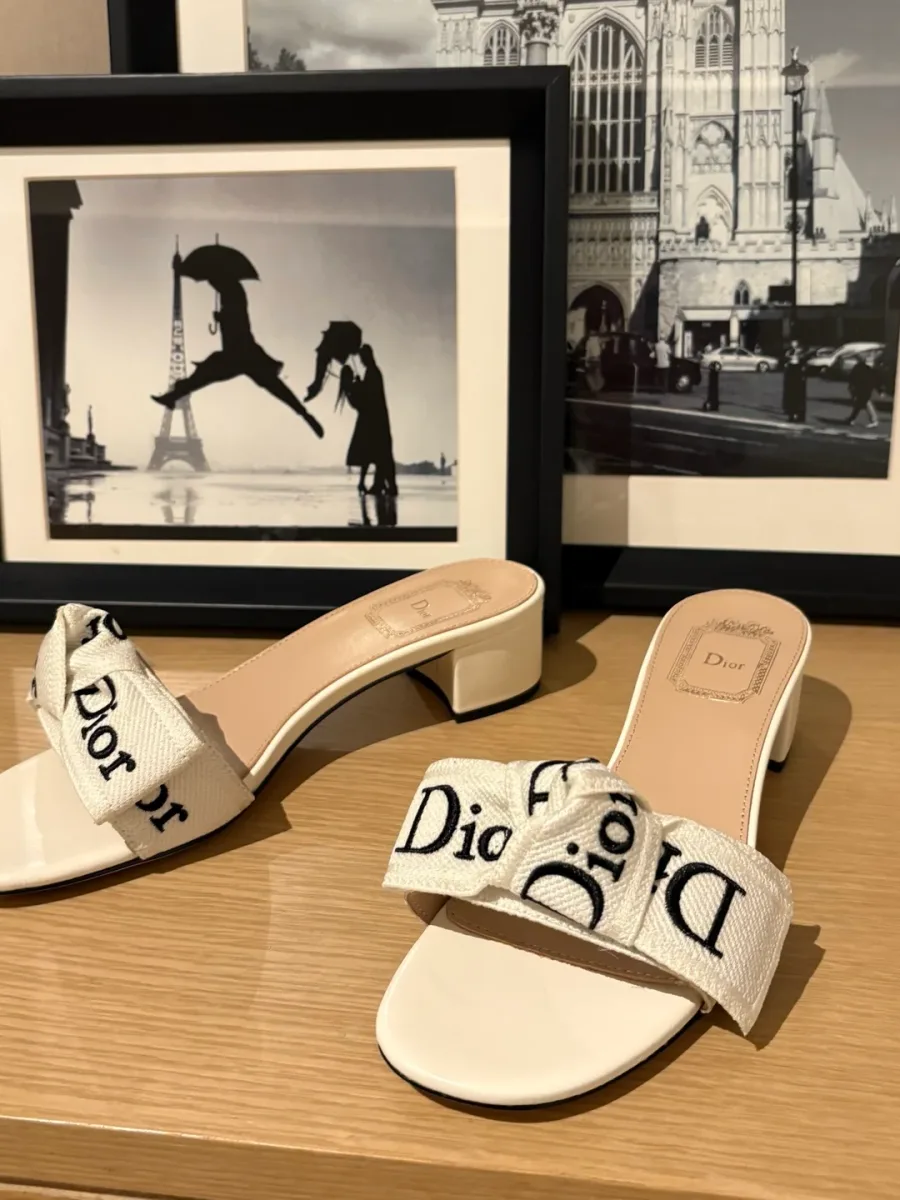 Белые шлёпанцы с бантом Dior на комоде перед картиной, ракурс спереди и сбоку
