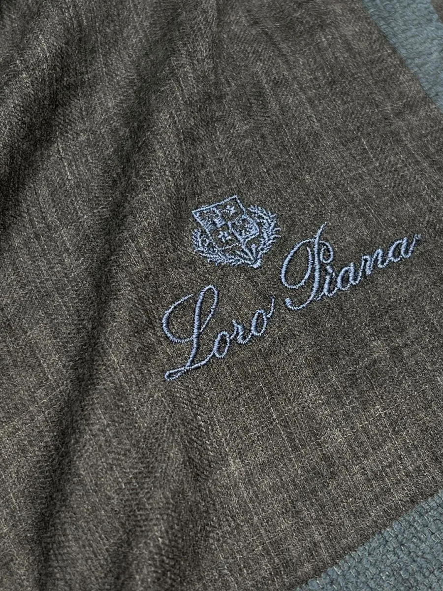 Крупный план вышитого логотипа Loro Piana голубой нитью на тёмно-сером полотне.