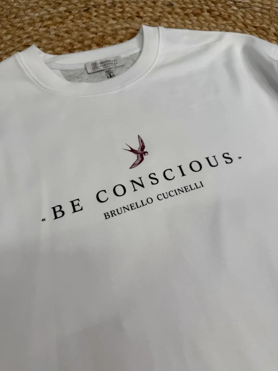 Вышитая ласточка и текстовый принт Be Conscious на груди белой футболки крупным планом